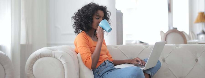 A woman sits on her couch to surf on the web and drink a cup of coffee.